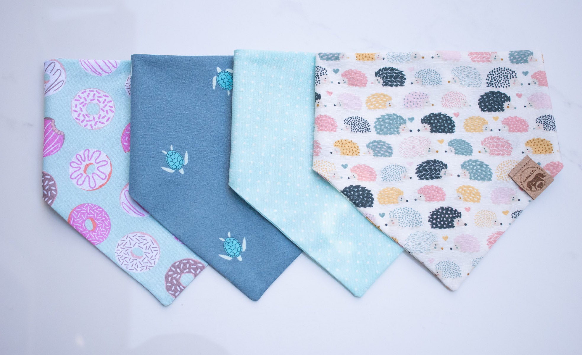 A photo of four pet bandanas. From left to right, featuring DoMutt, Turtle Reef, Dip n Dots and Hedgehog Love