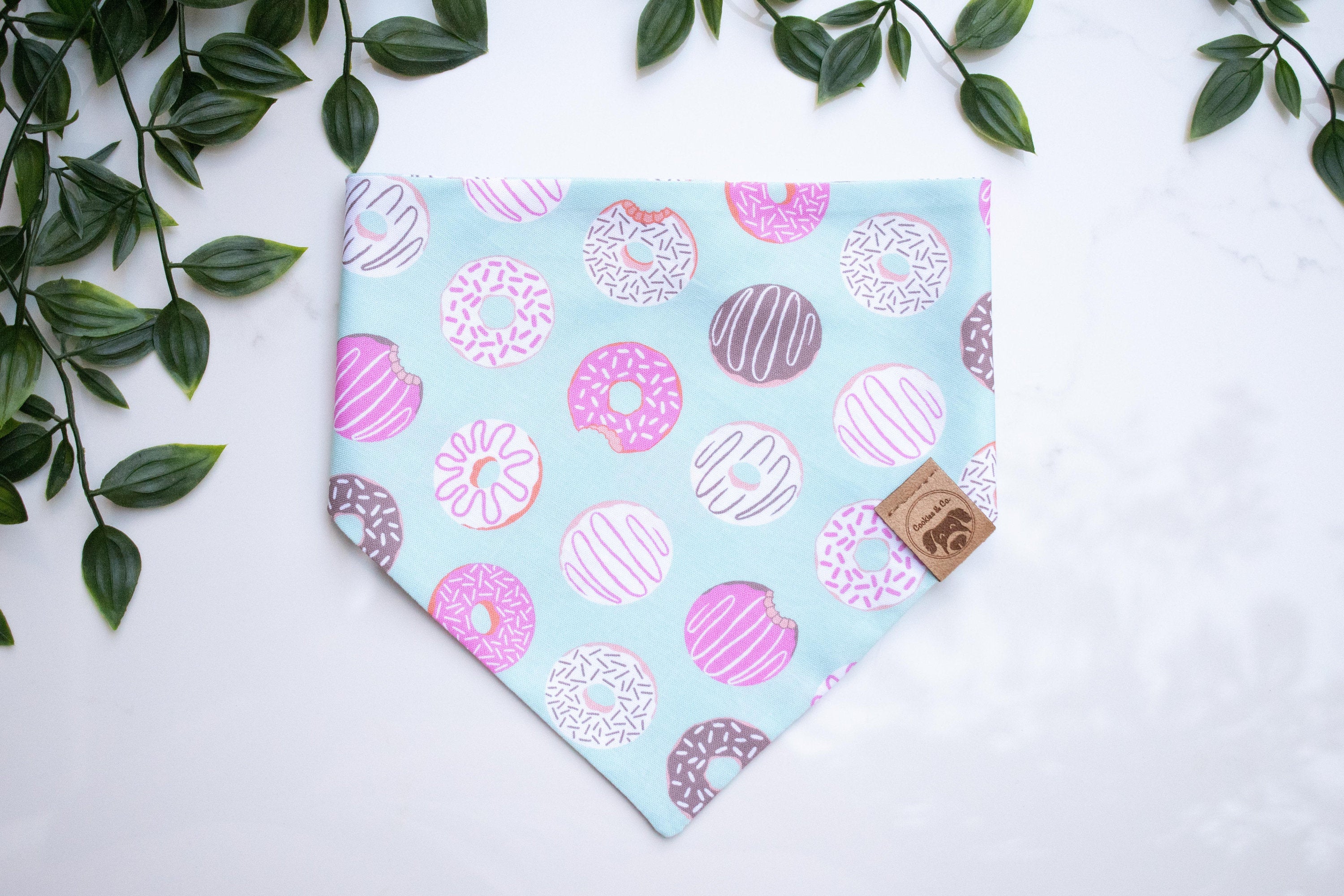 DoMutts bandana print, featuring all sorts of donuts such as jelly filled donuts, chocolate glazed donuts with white sprinkles, arranged row by row diagonally, on a light blue fabric